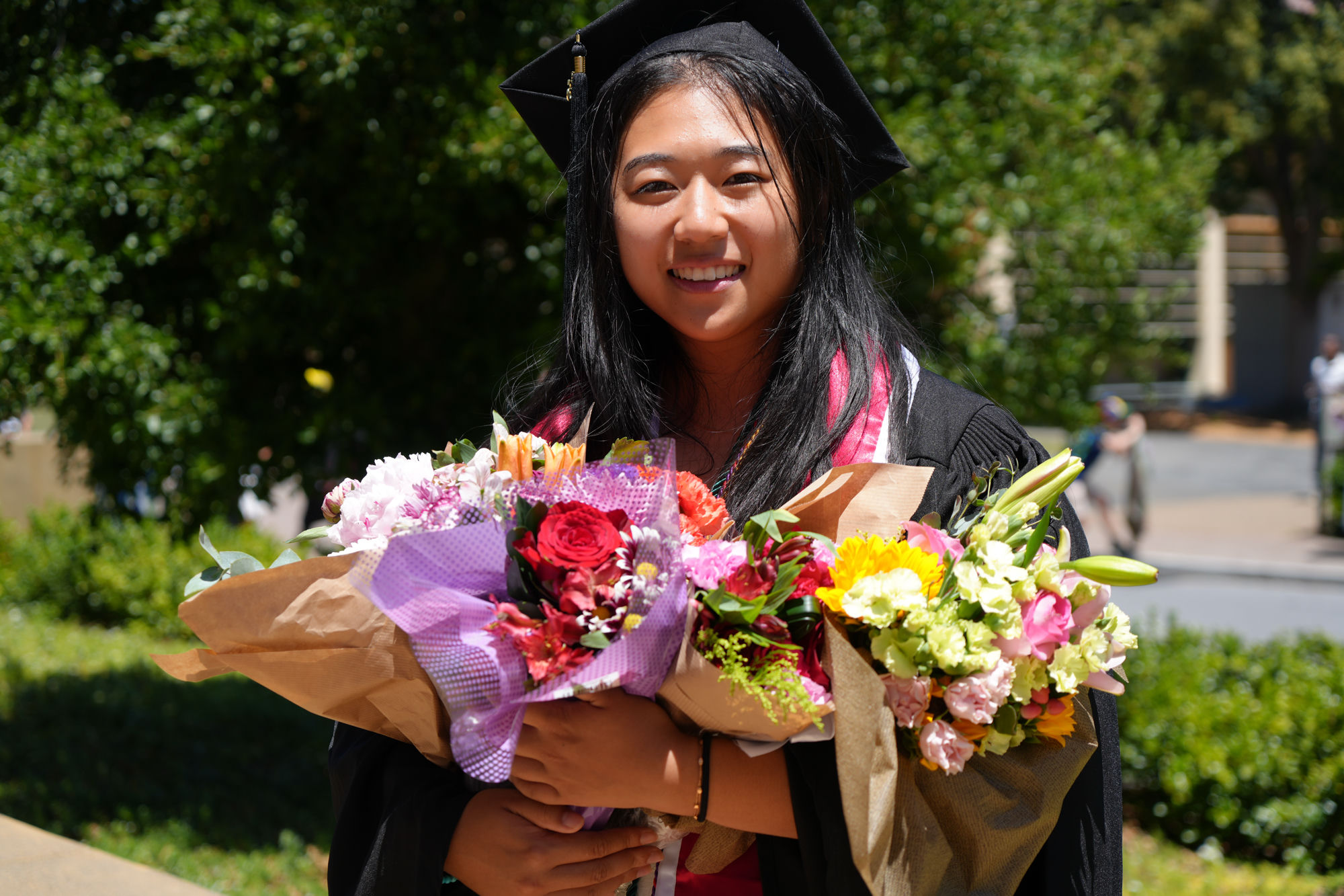 Artemis Xu in cap and gown holding bouquets of flowers on graduation day